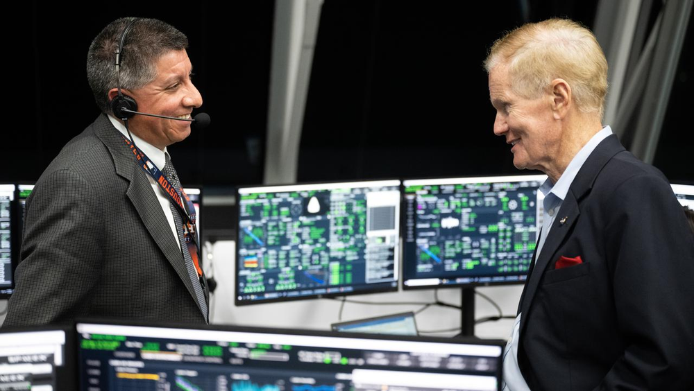 Two people in suits smile at each other standing in front of monitors on a desk with a black background.